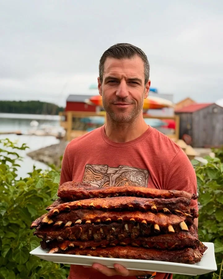 A person holding a large platter stacked with barbecue ribs outdoors, with a scenic background.