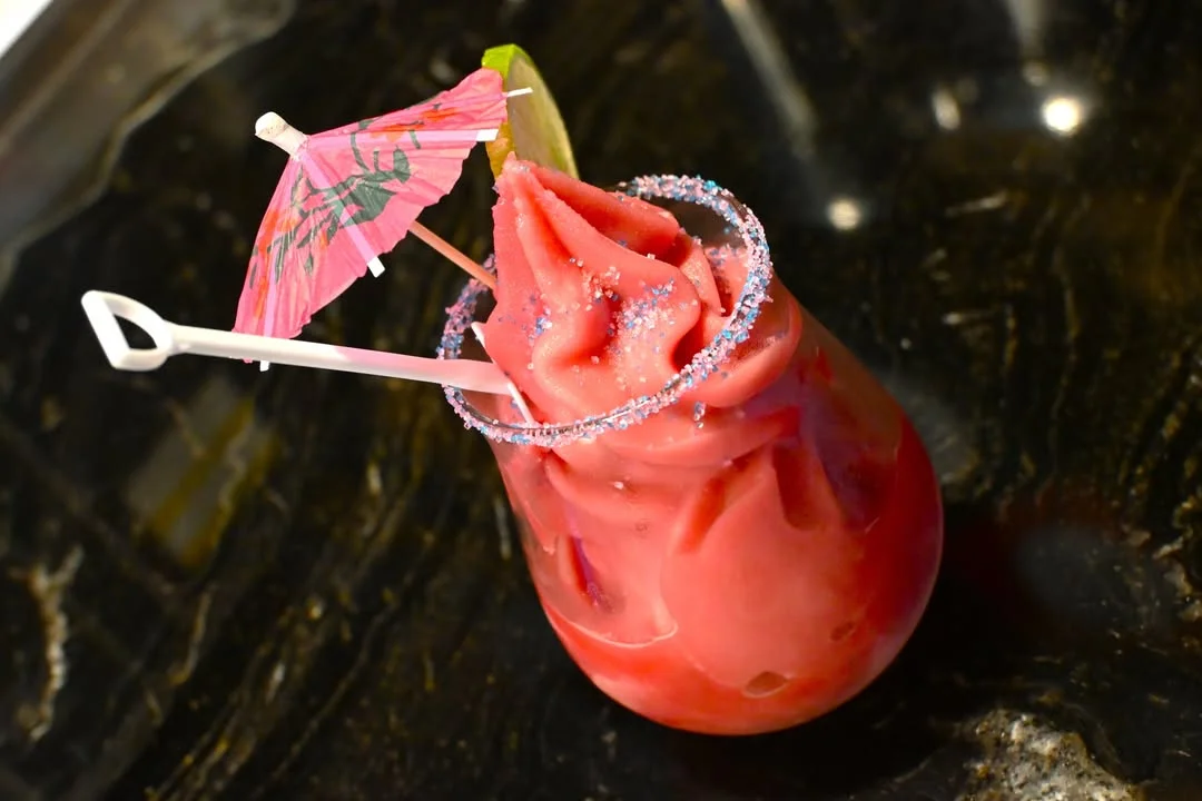A vibrant frozen drink with a pink umbrella and sugar rim, topped with a swirled ice cream texture.