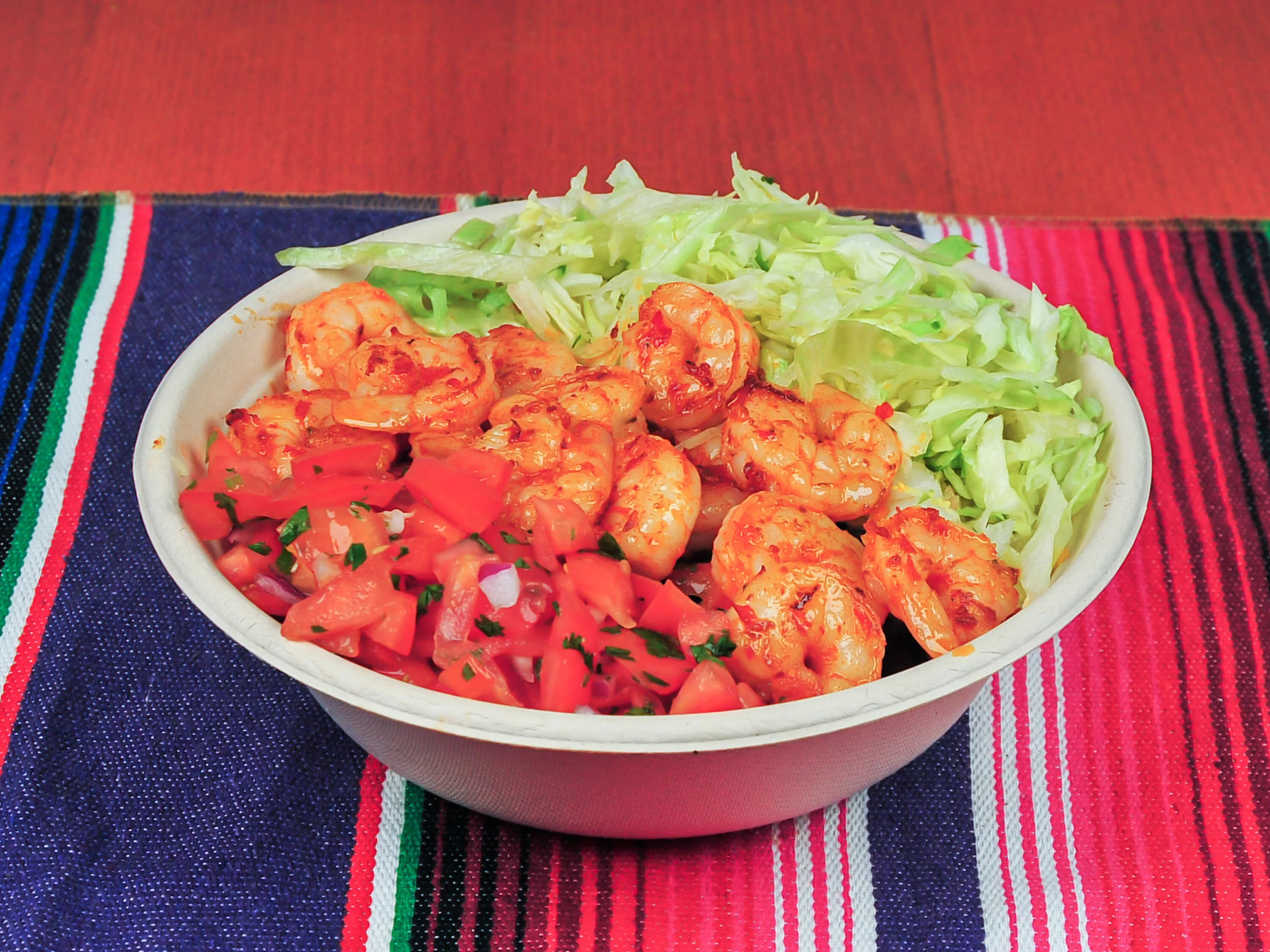 A bowl filled with sautéed shrimp, diced tomatoes mixed with onions and herbs, accompanied by a serving of shredded lettuce. The bowl is placed on a colorful striped tablecloth.