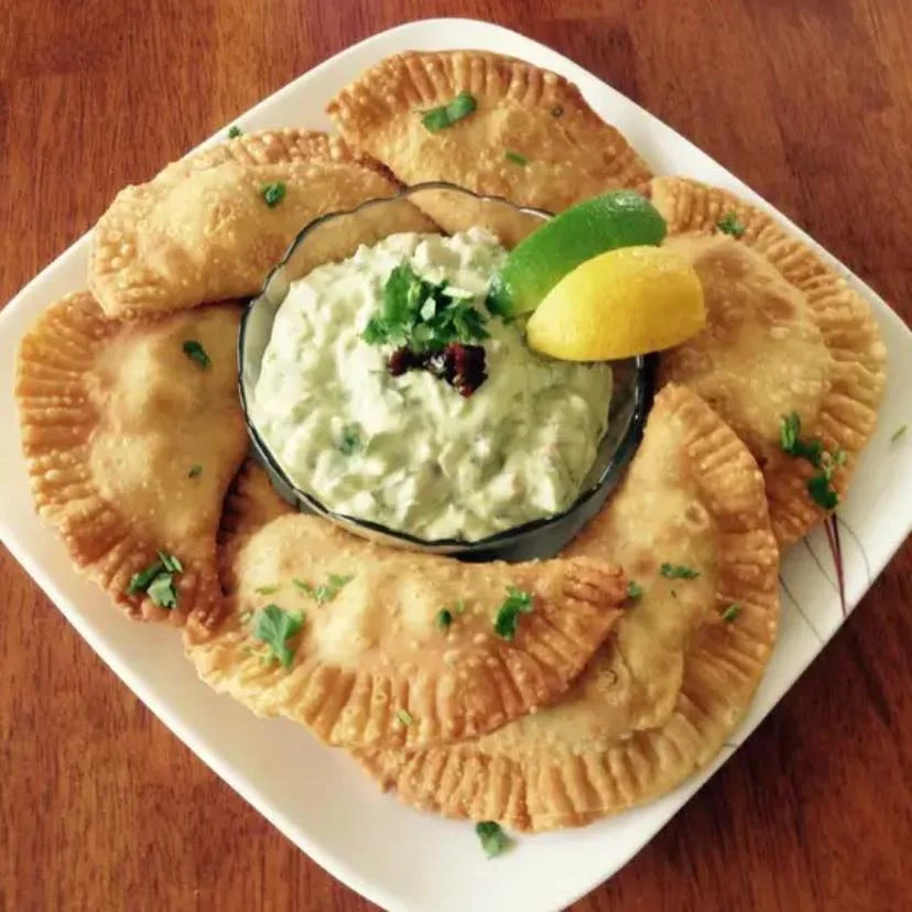 A plate featuring fried pastries arranged around a bowl of creamy dip, garnished with lime and lemon.