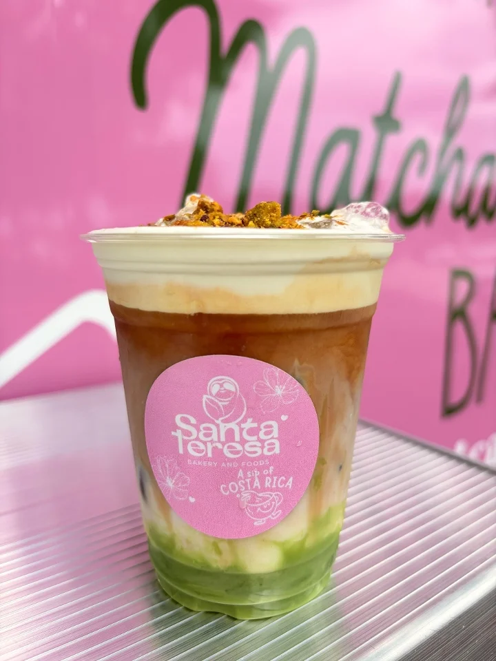 A colorful drink with layered matcha and milk, topped with cream and crunchy garnish in a clear cup.