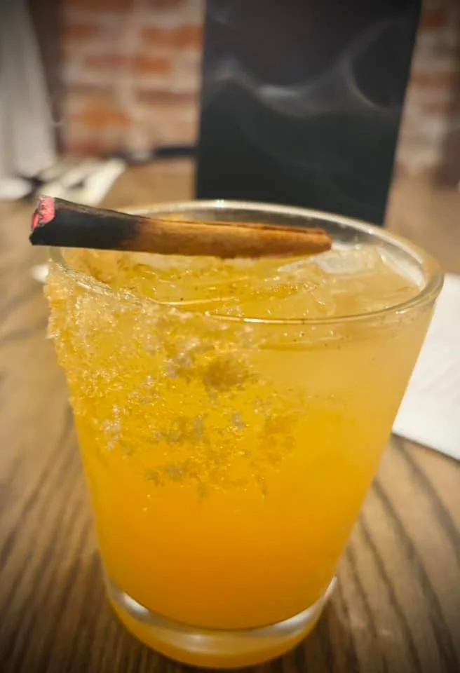A cocktail in a glass with ice, garnished with a burnt cinnamon stick, showcasing a vibrant orange color.