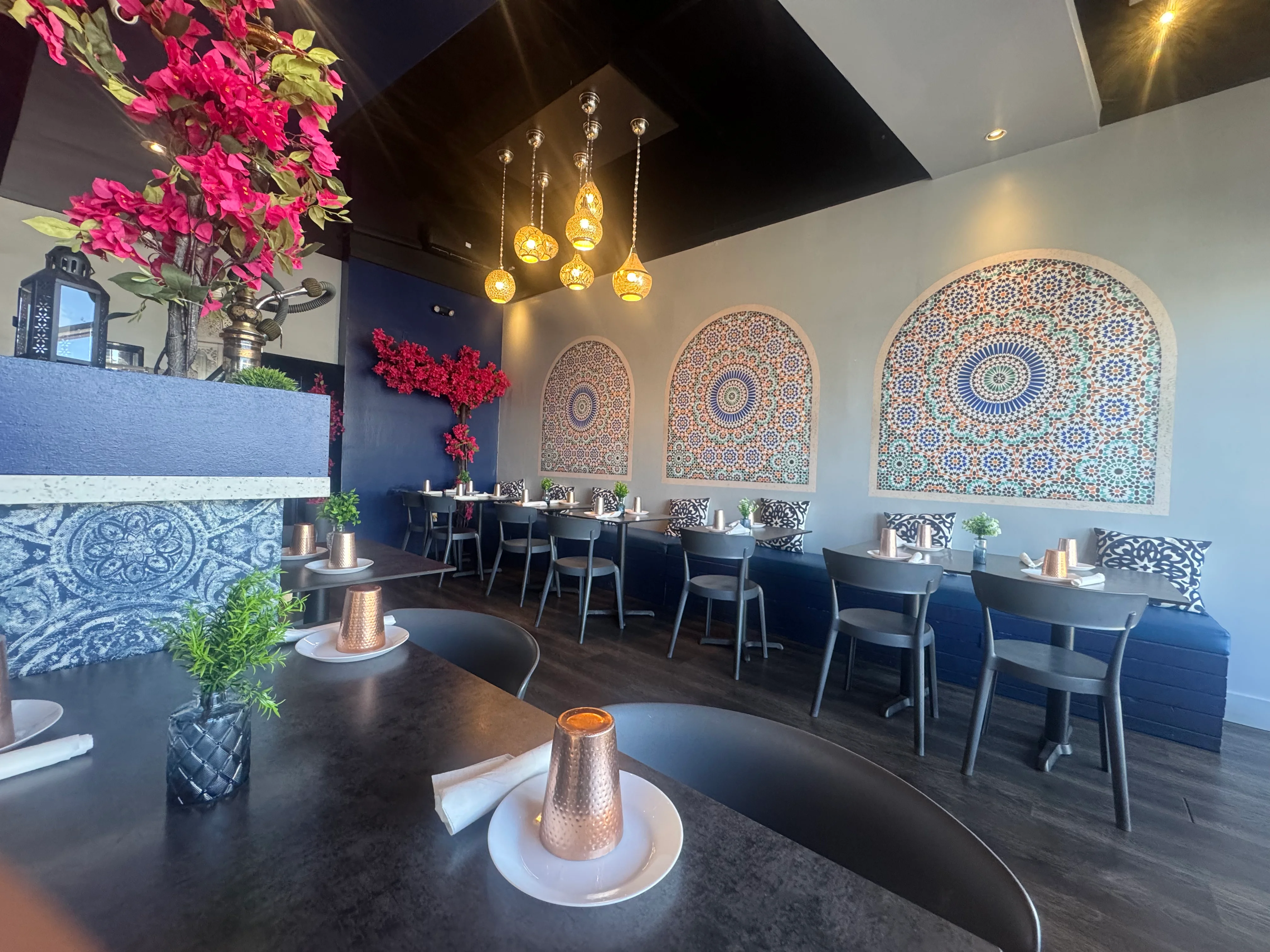 Stylish café interior with wooden floors and modern decor. A patterned counter with plants, cozy chairs, and ambient lighting creates a welcoming atmosphere.