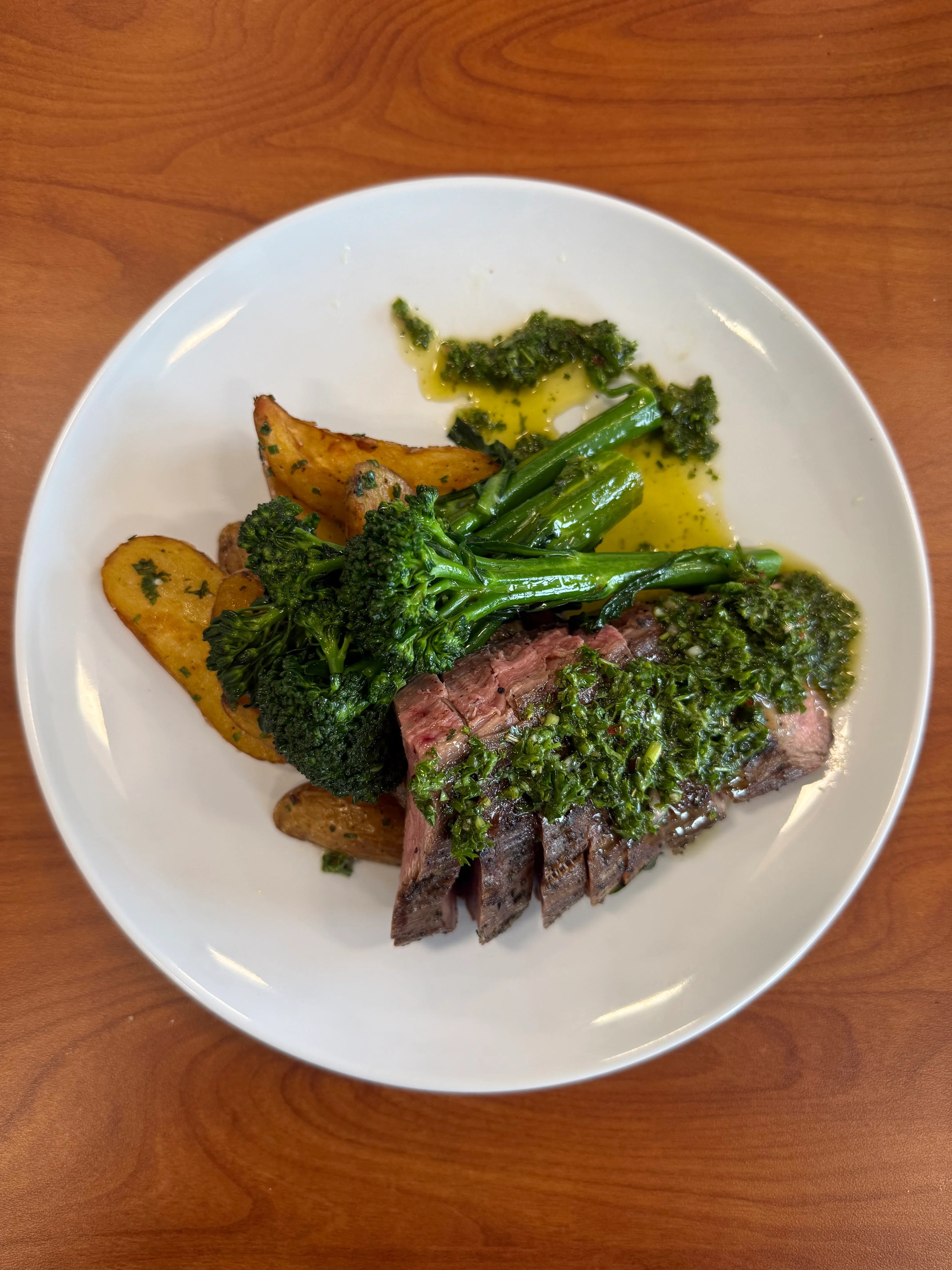 Sliced steak with chimichurri, roasted potatoes, and broccolini on a plate.