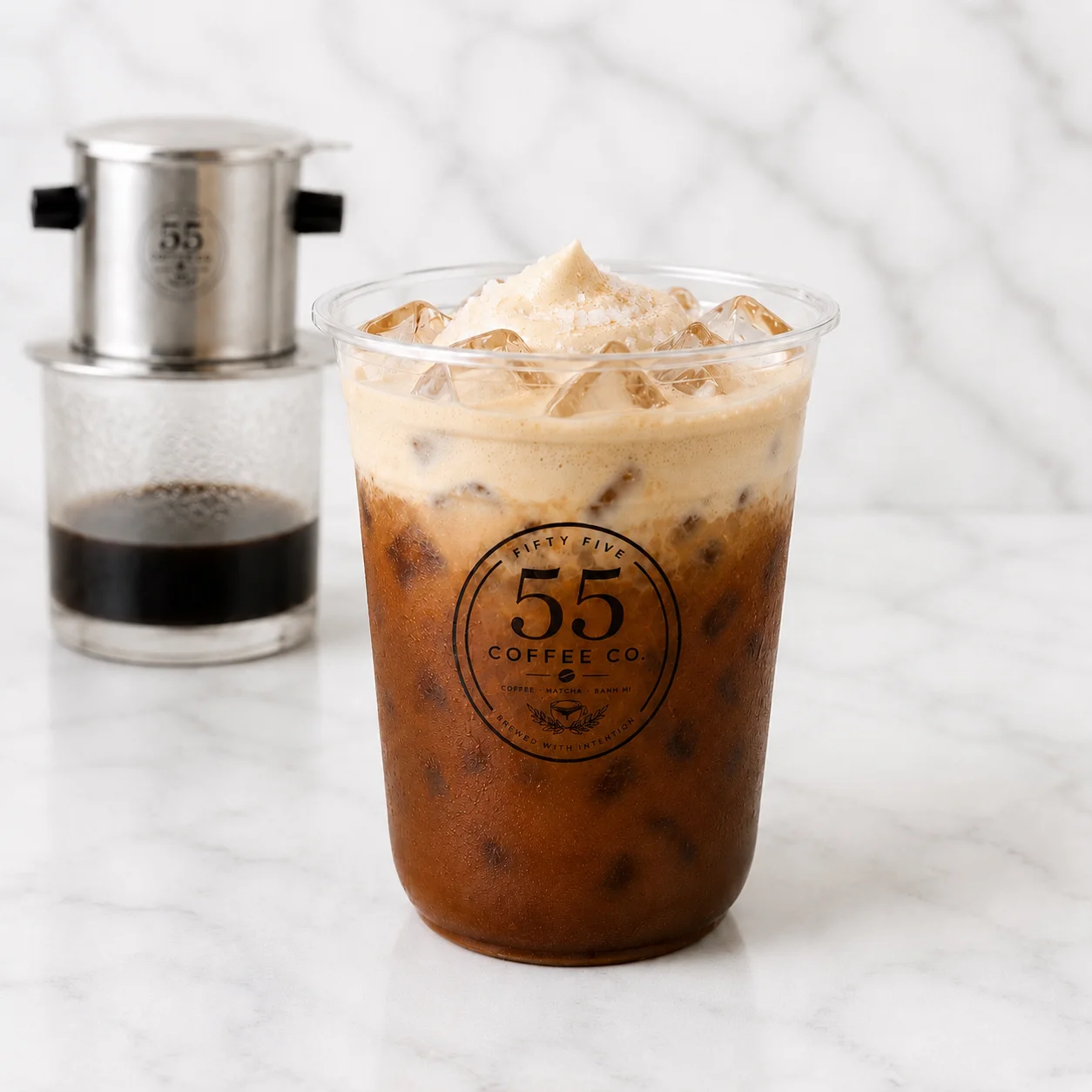 A refreshing iced coffee topped with a frothy layer, served in a clear cup with a logo. A traditional coffee filter sits in the background.