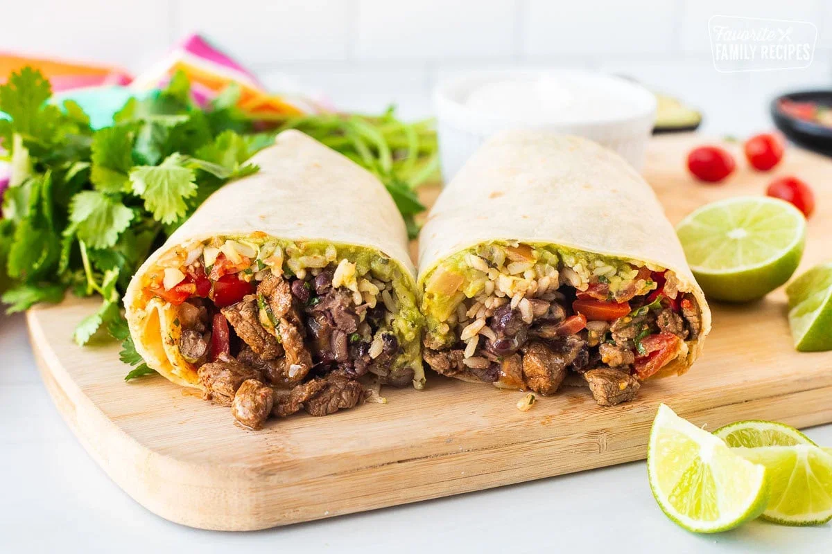 Two halves of a burrito filled with meat, rice, and fresh vegetables, placed on a wooden cutting board with lime slices and herbs nearby.