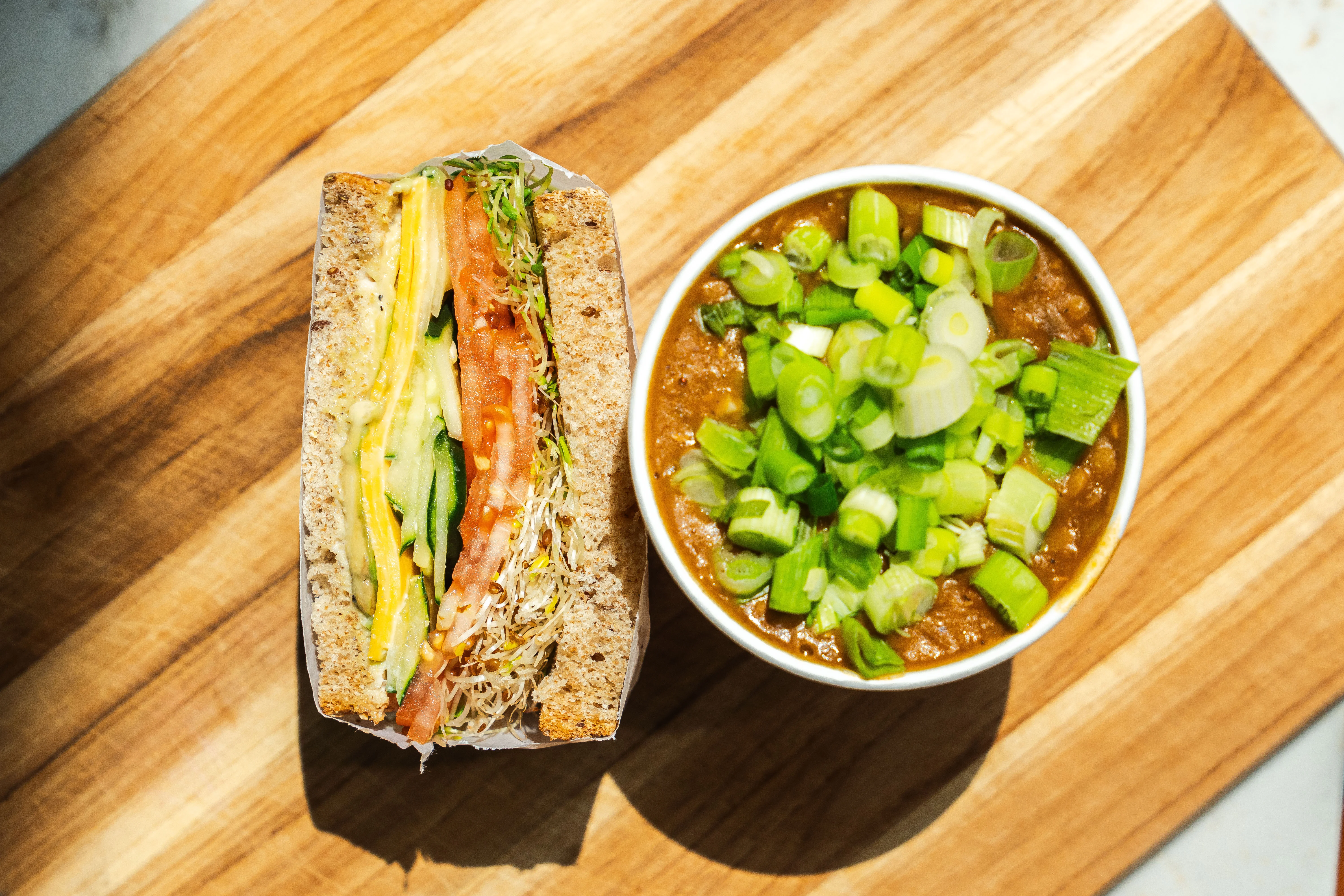 An overhead view of a sandwich stacked with vegetables, served alongside a bowl of soup garnished with green onions. The wooden surface adds a rustic touch.