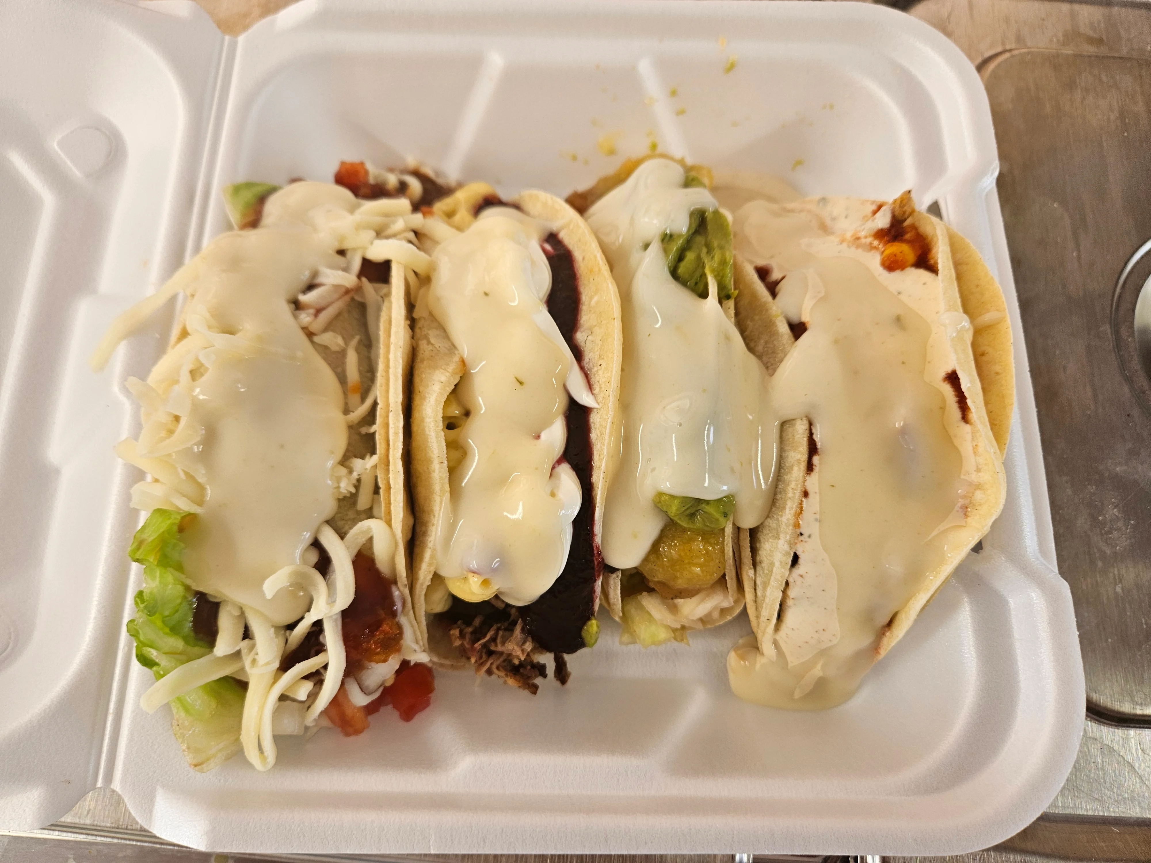 A takeout container filled with four tacos, each topped with various ingredients and a creamy sauce. The tacos are placed side by side for display.