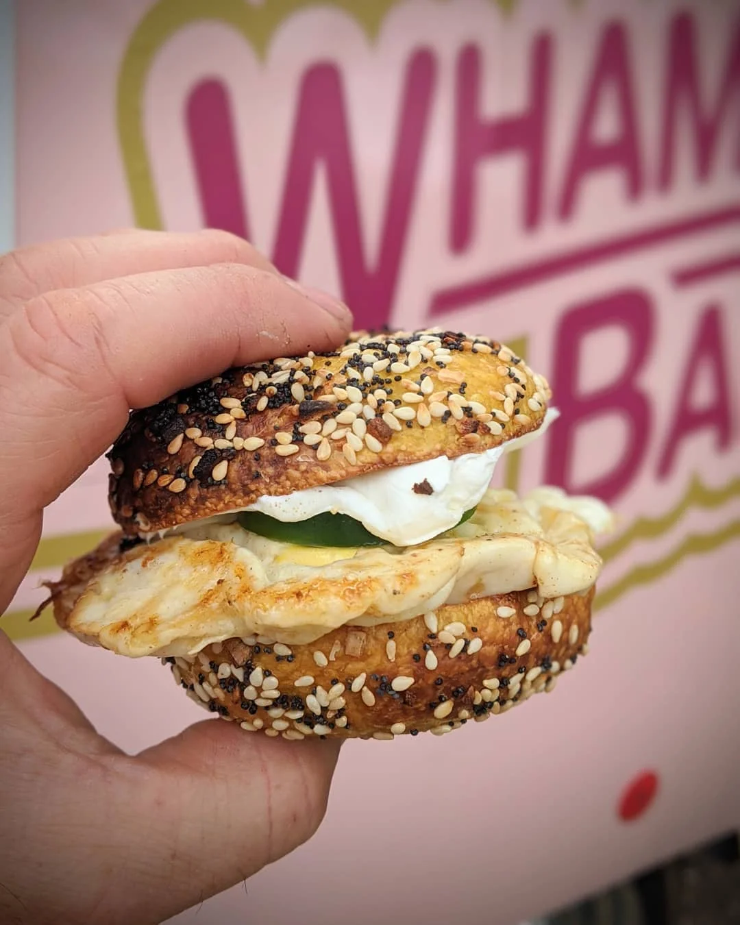 A hand holding a sesame seed bagel sandwich filled with egg, vegetables, and sauce, set against a colorful background.
