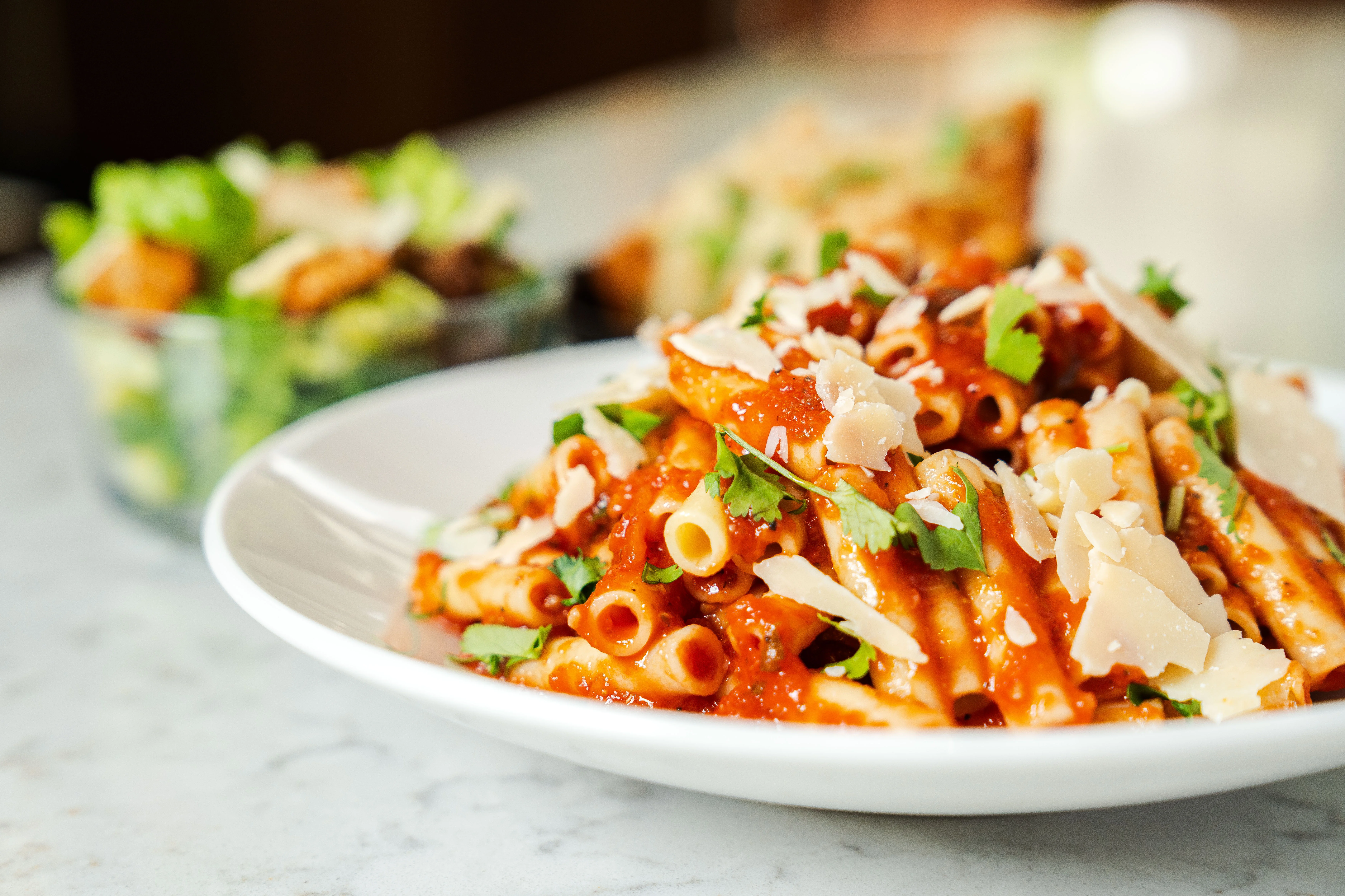 A plate of pasta topped with marinara sauce, garnished with cilantro and shaved parmesan cheese. A small salad is served in the background.
