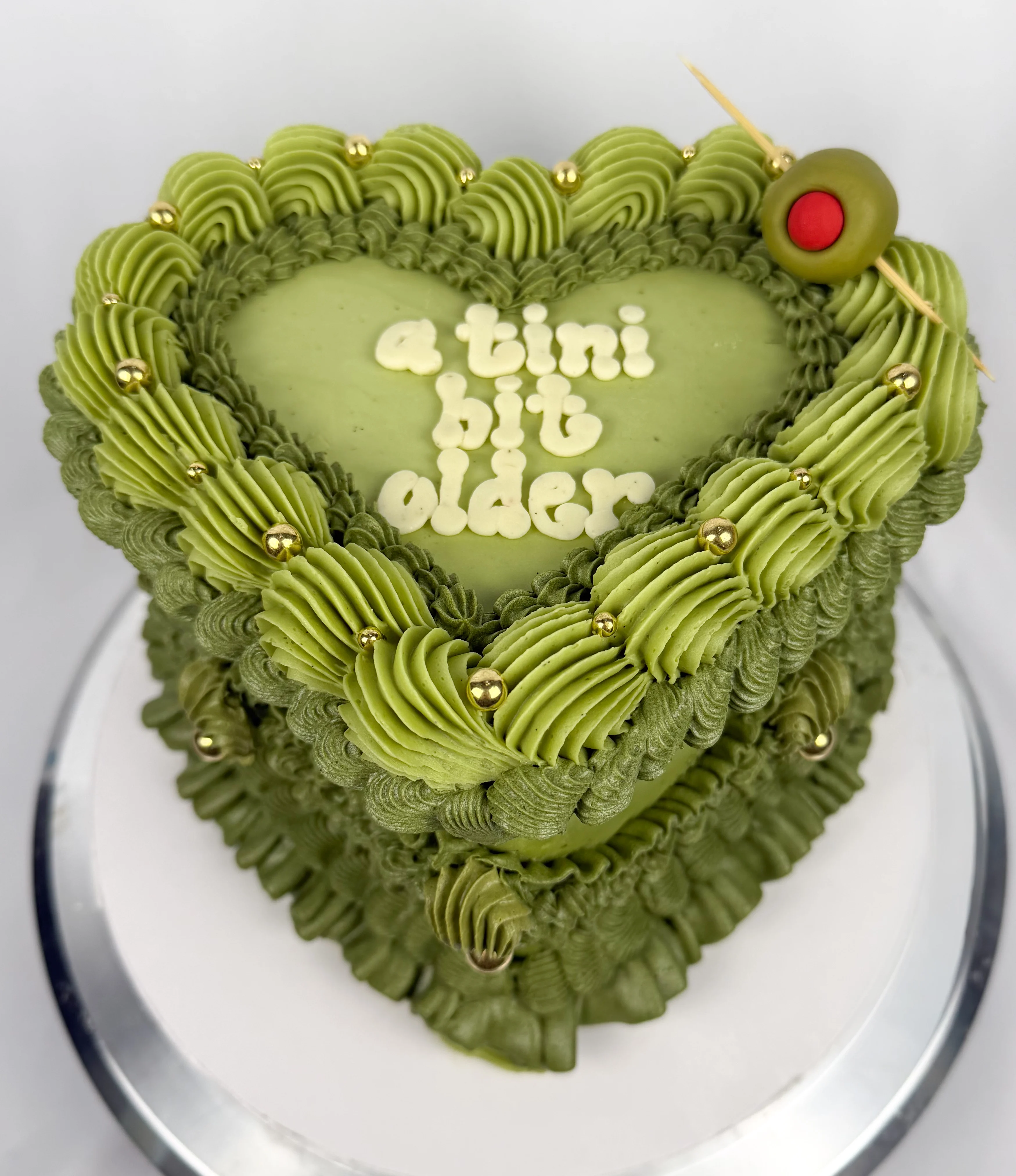 A unique green heart-shaped cake with a layered design, adorned with swirled frosting, gold accents, and a decorative olive on top.