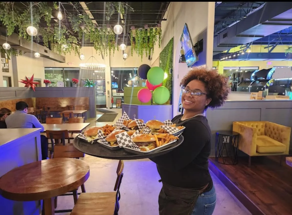 Server carrying a tray of burgers and fries in a vibrant restaurant setting.