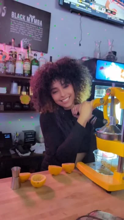 A bartender smiling while preparing fresh drinks, with spirits in the background and colorful lighting enhancing the atmosphere.