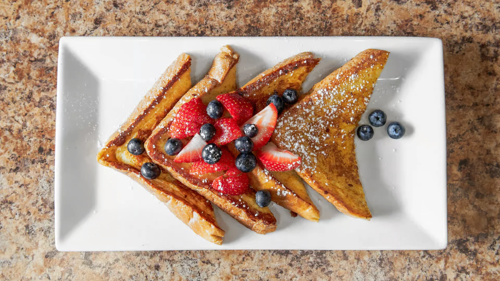 A plate of golden-brown French toast from El Ranchito Poblano, topped with powdered sugar, fresh strawberries, and blueberries.