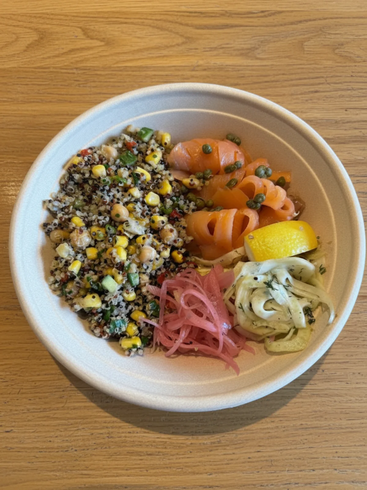 A bowl featuring quinoa salad with corn, chickpeas, and diced vegetables, accompanied by salmon slices and pickled onions, garnished with lemon.