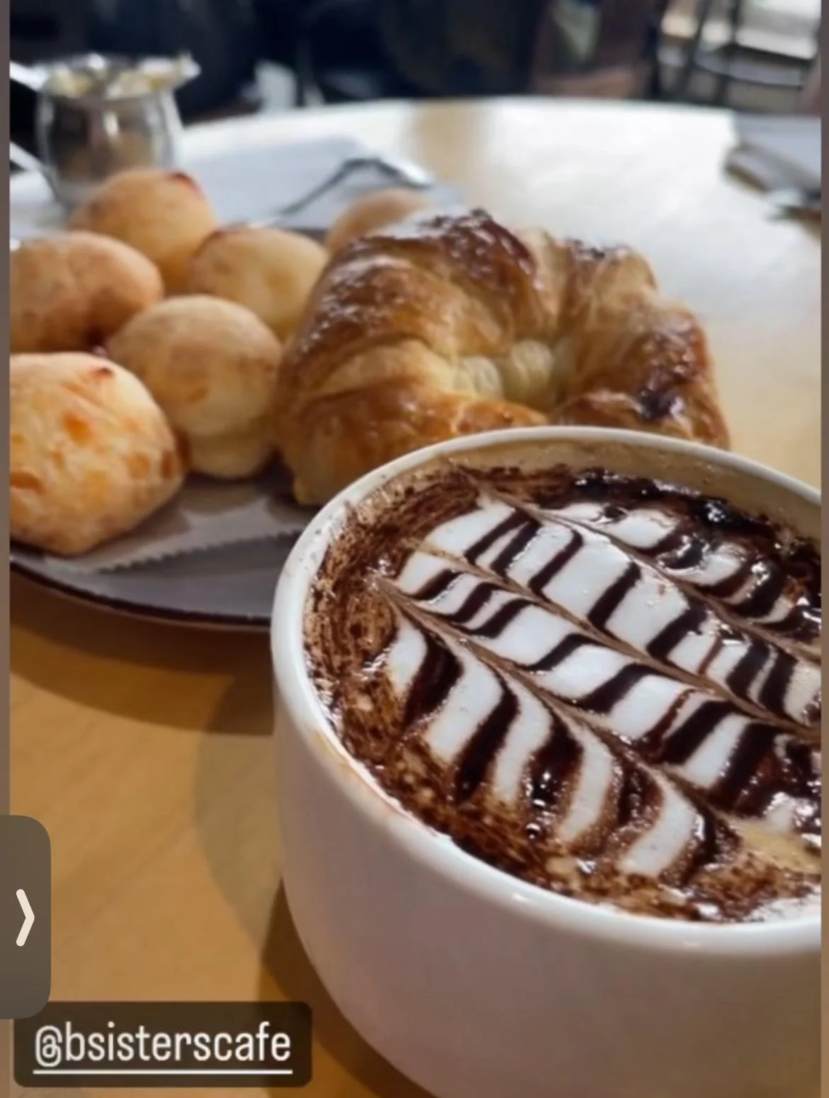 A cup of chocolate mocha topped with a decorative foam design, accompanied by various baked goods including a croissant and cheese bread rolls on a plate.