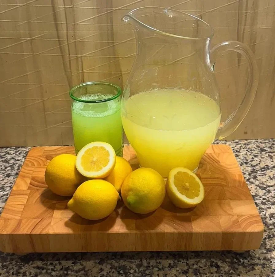A wooden board holds fresh lemons and a pitcher of lemonade, with a glass of green drink nearby.