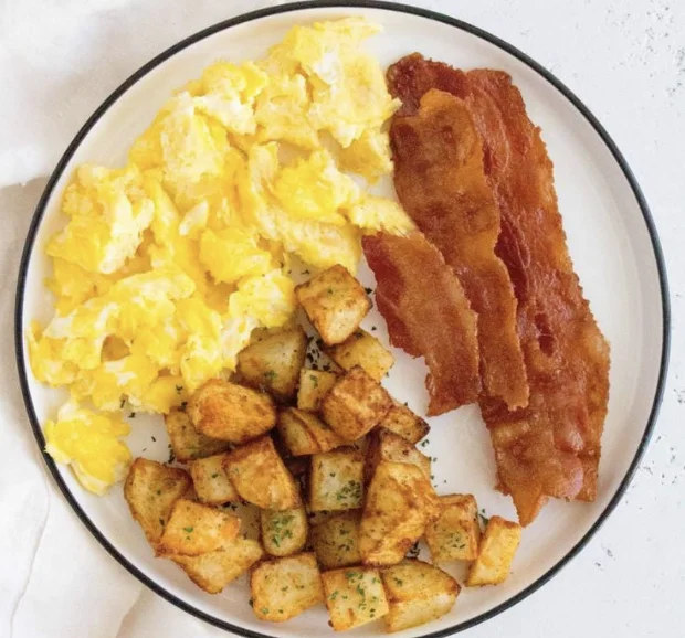 A breakfast plate featuring scrambled eggs, crispy bacon strips, and golden fried potatoes, arranged neatly.