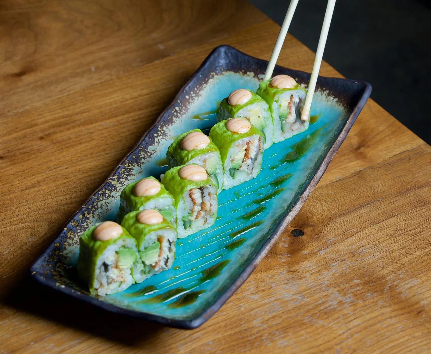 Sushi rolls wrapped in bright green soy paper or avocado, topped with spicy mayo and served on a turquoise rectangular plate being picked up with chopsticks.