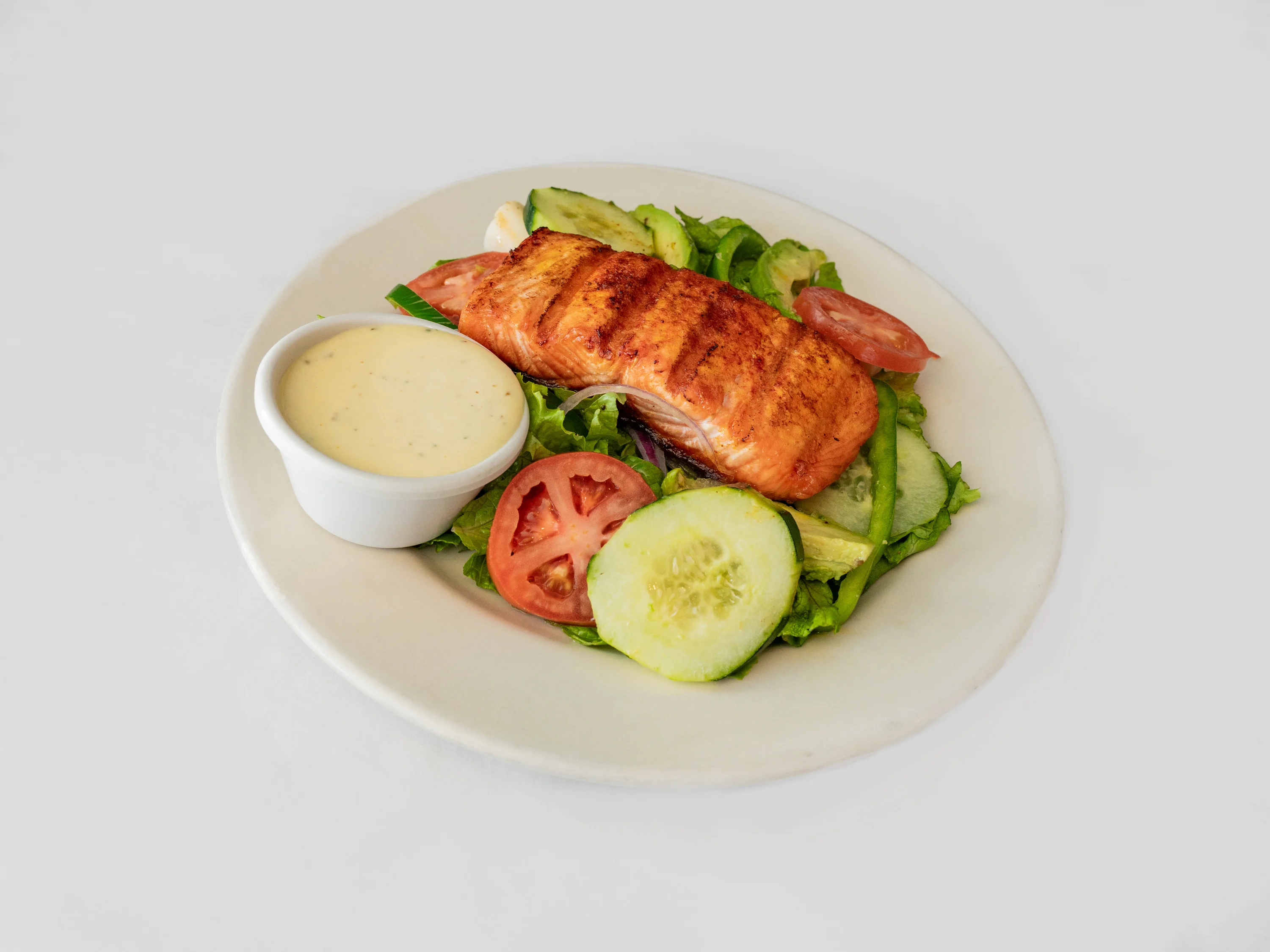 A grilled salmon fillet on a bed of fresh greens, accompanied by slices of cucumber and tomato, and served with a creamy dressing.