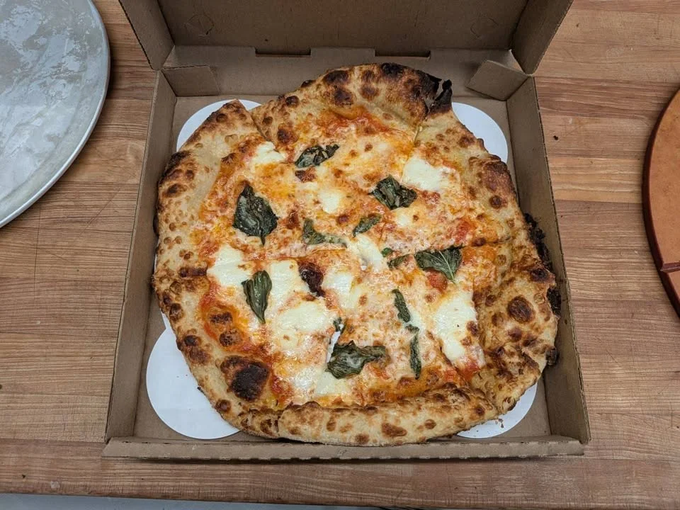 A Margherita pizza in a brown delivery box, topped with fresh basil and mozzarella cheese.