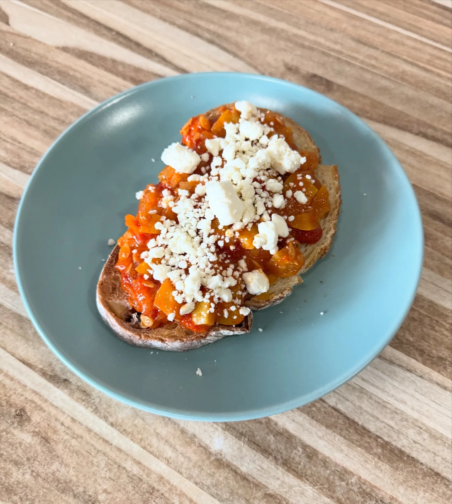 A slice of bread topped with a savory sauce made from chopped vegetables and crumbled feta cheese, served on a light blue plate.