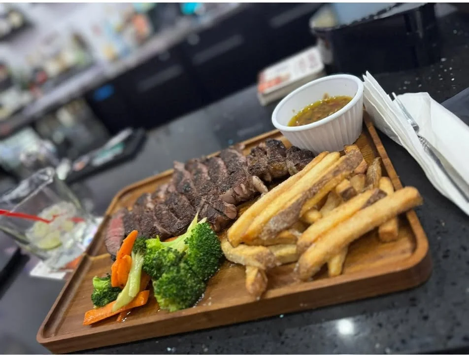 A wooden platter featuring sliced steak, golden fries, steamed broccoli, and a small bowl of dipping sauce.