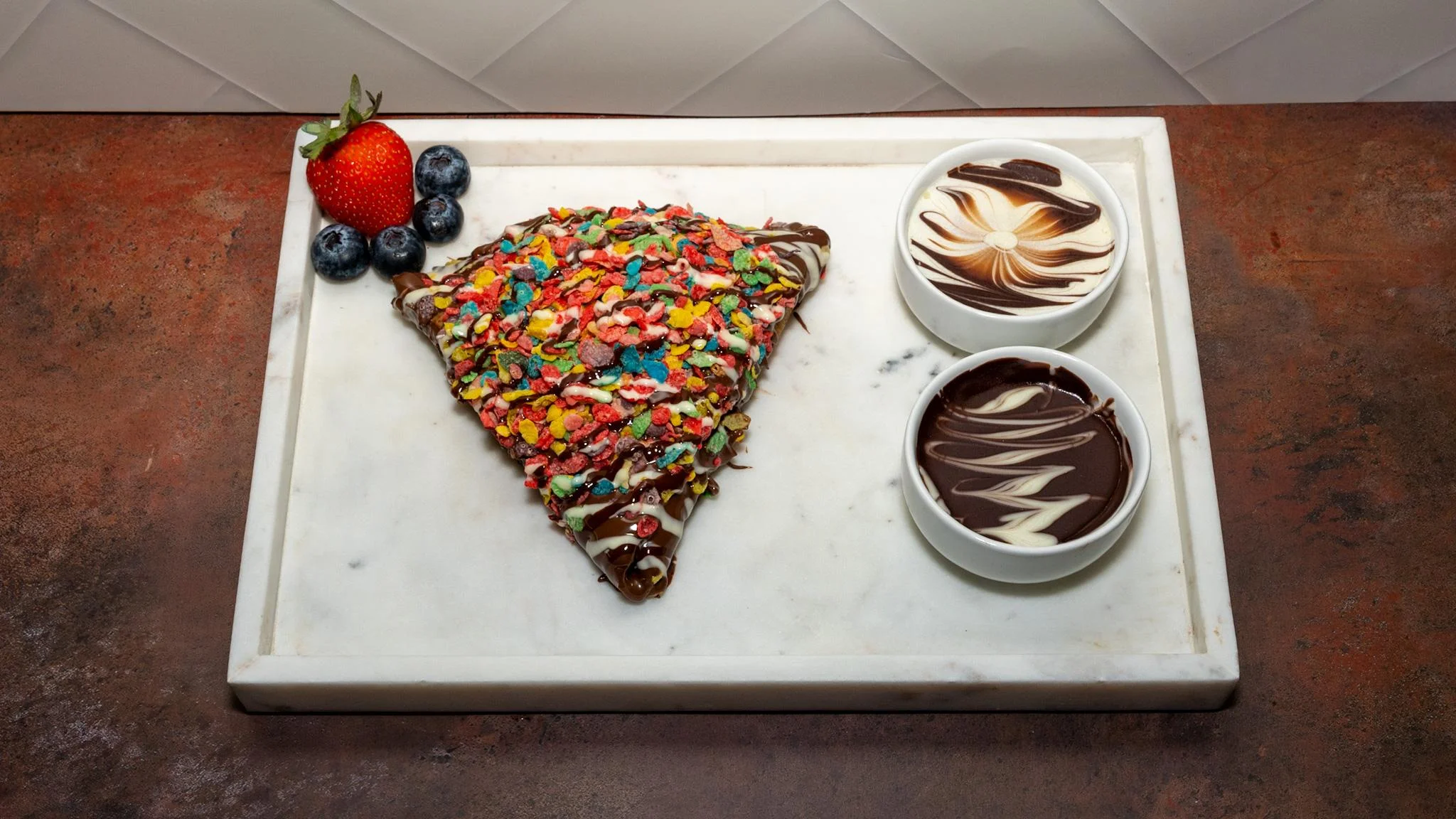 A chocolate-covered crepe topped with colorful cereal and accompanied by bowls of chocolate sauces. Fresh strawberries and blueberries are also present.