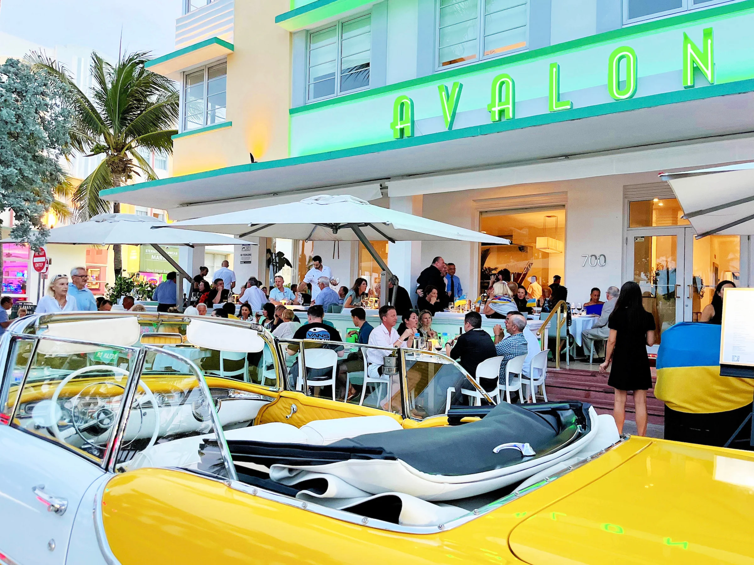 A vibrant Art Deco building with palm trees and an American flag, featuring an outdoor dining area.