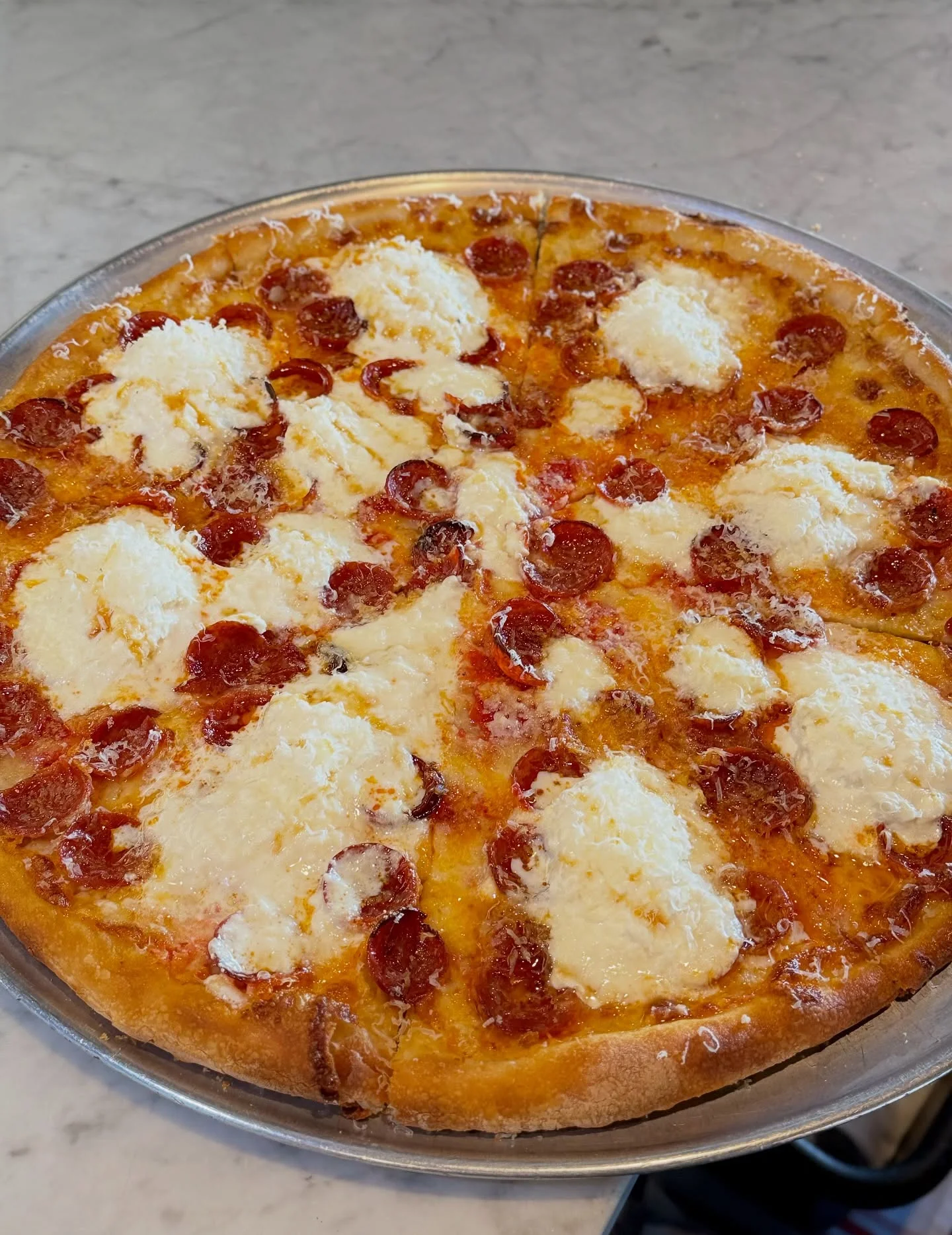 A round pizza topped with pepperoni slices and fluffy dollops of ricotta cheese, presented on a metallic tray against a marble background.