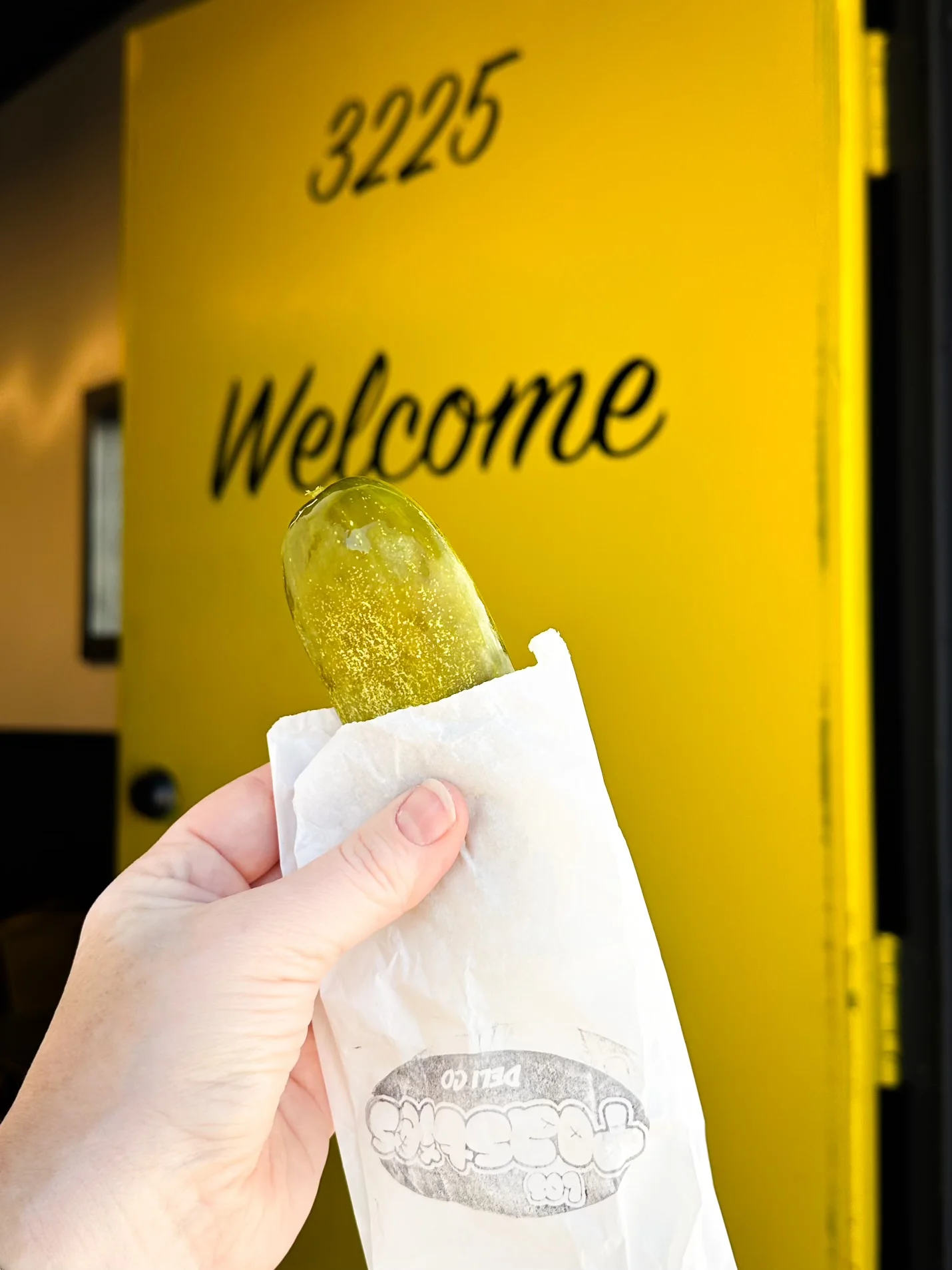 Handheld pickle wrapped in paper at a vibrant restaurant entrance