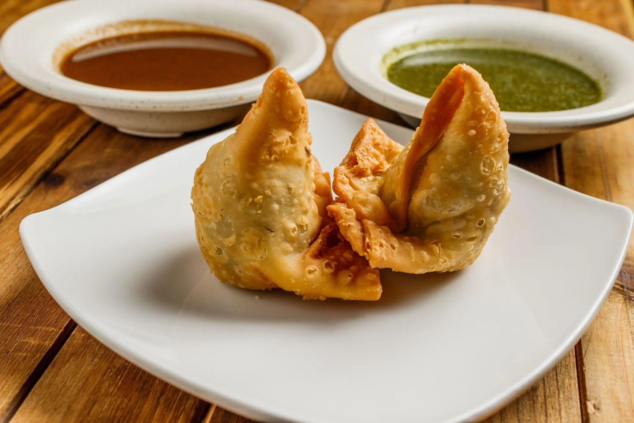 Two crispy fried pastries shaped like triangles, served on a white plate with two different dipping sauces in the background.
