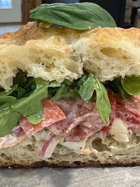 A close-up of a sandwich featuring layers of meats, greens, and a flaky bread. The sandwich includes salami, fresh basil, and arugula.