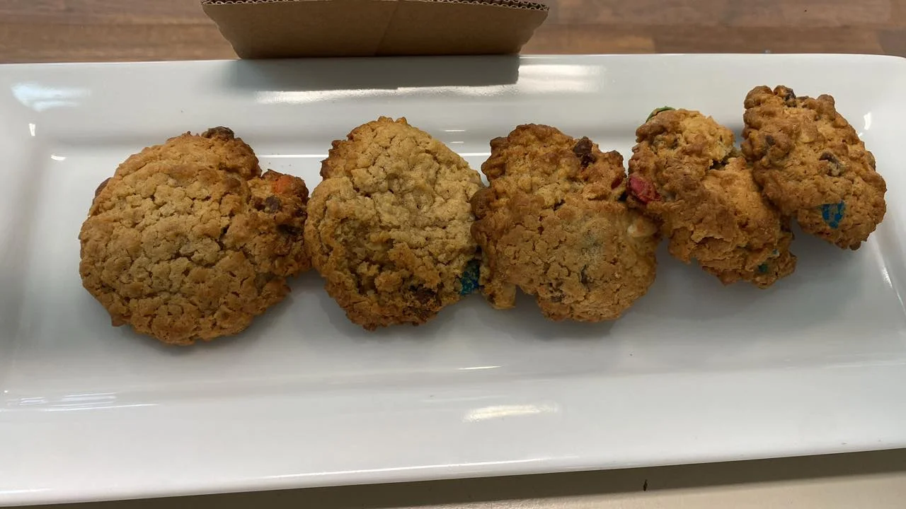Five oatmeal cookies displayed on a long white platter, showcasing a golden-brown exterior with visible pieces of chocolate and colorful bits.