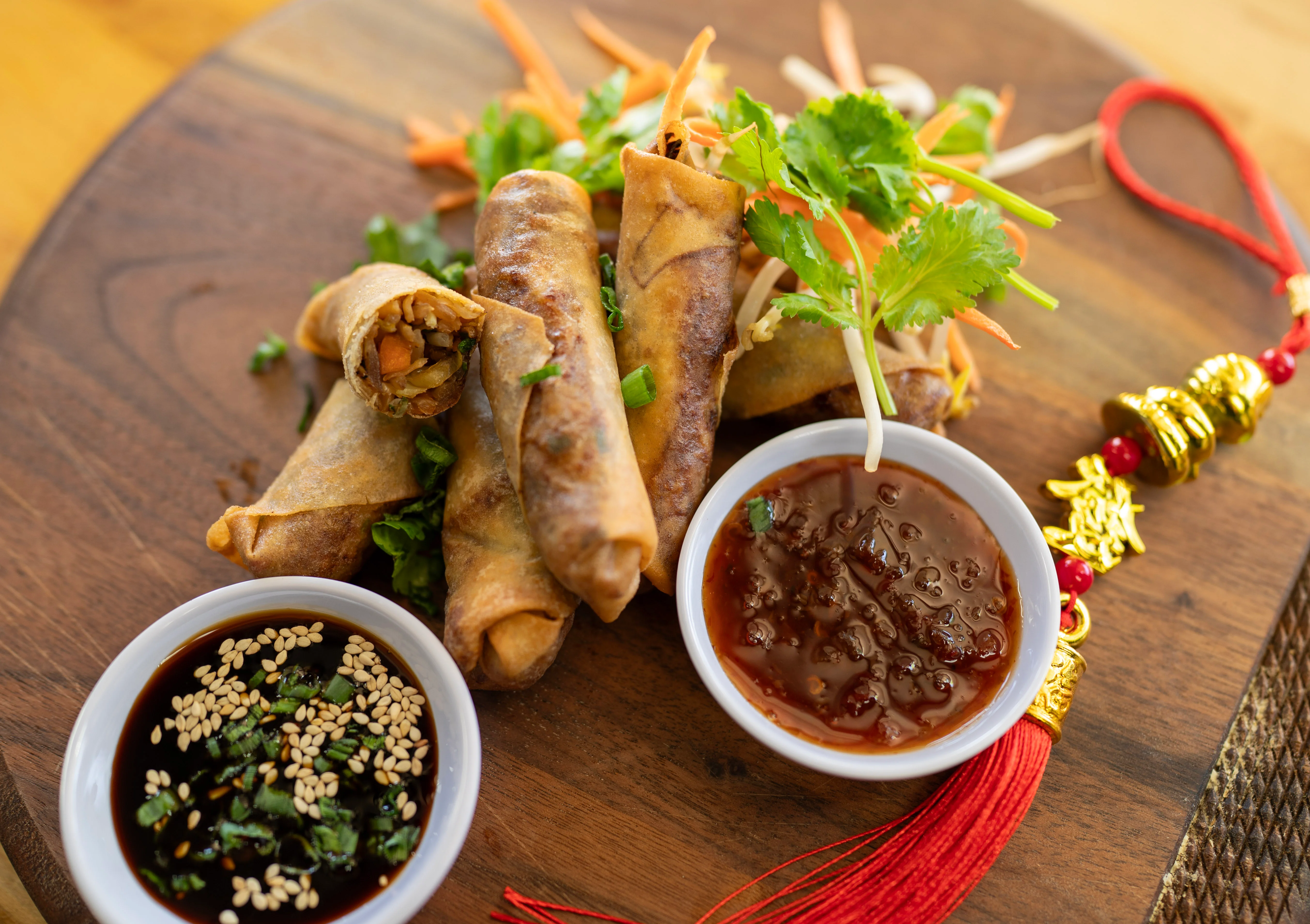 A wooden platter featuring crispy spring rolls accompanied by dipping sauces and fresh vegetables.