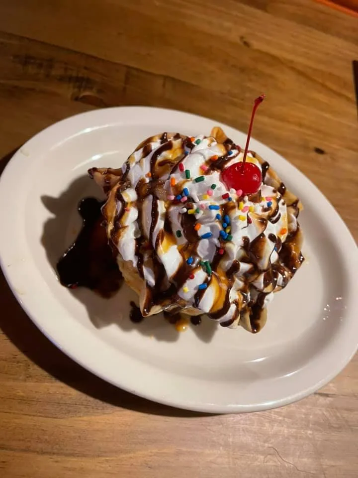 A dessert featuring a scoop of ice cream topped with whipped cream, chocolate syrup, colorful sprinkles, and a cherry.