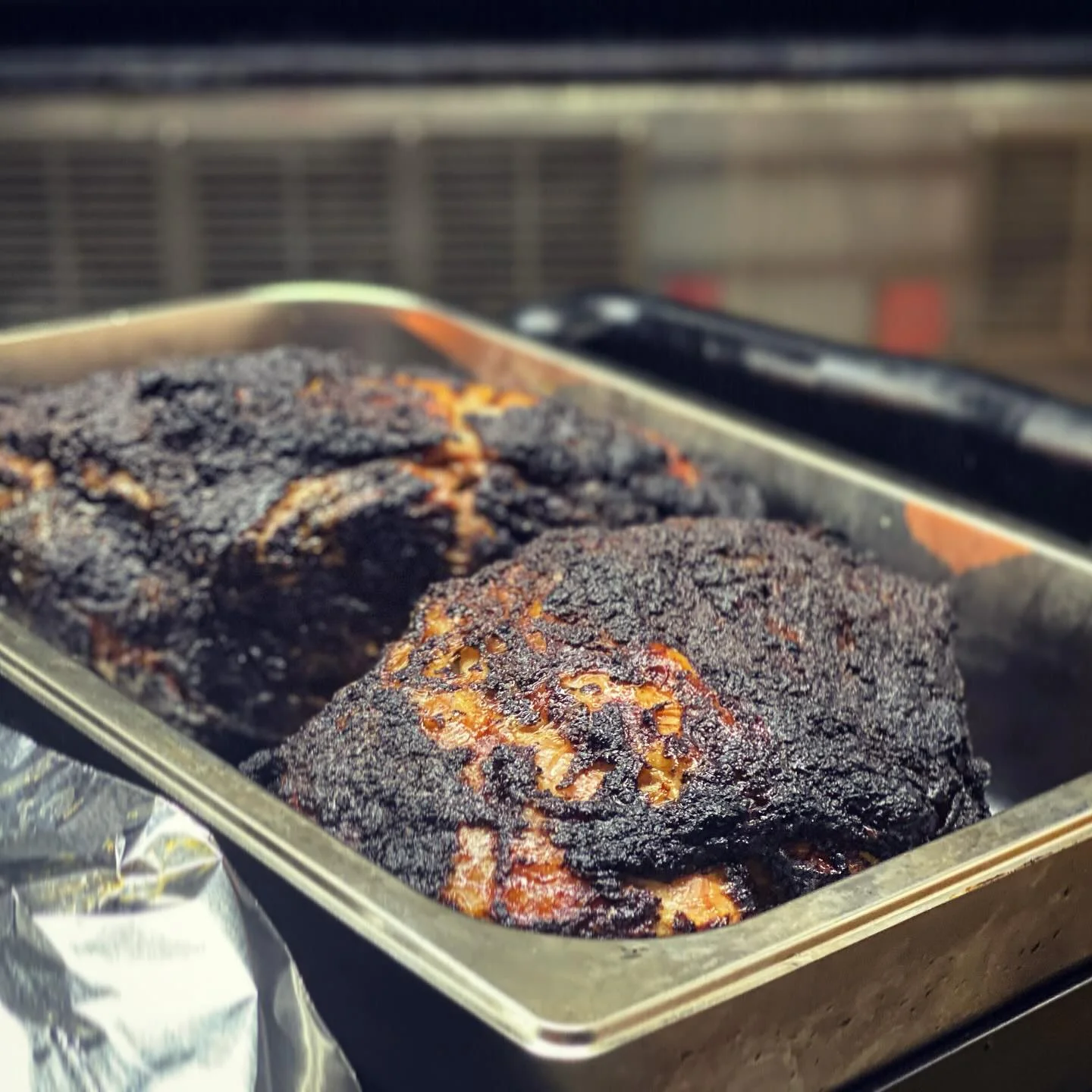 Two pieces of smoked meat in a silver tray, showcasing a dark, charred exterior, indicating a slow-cooked preparation method.