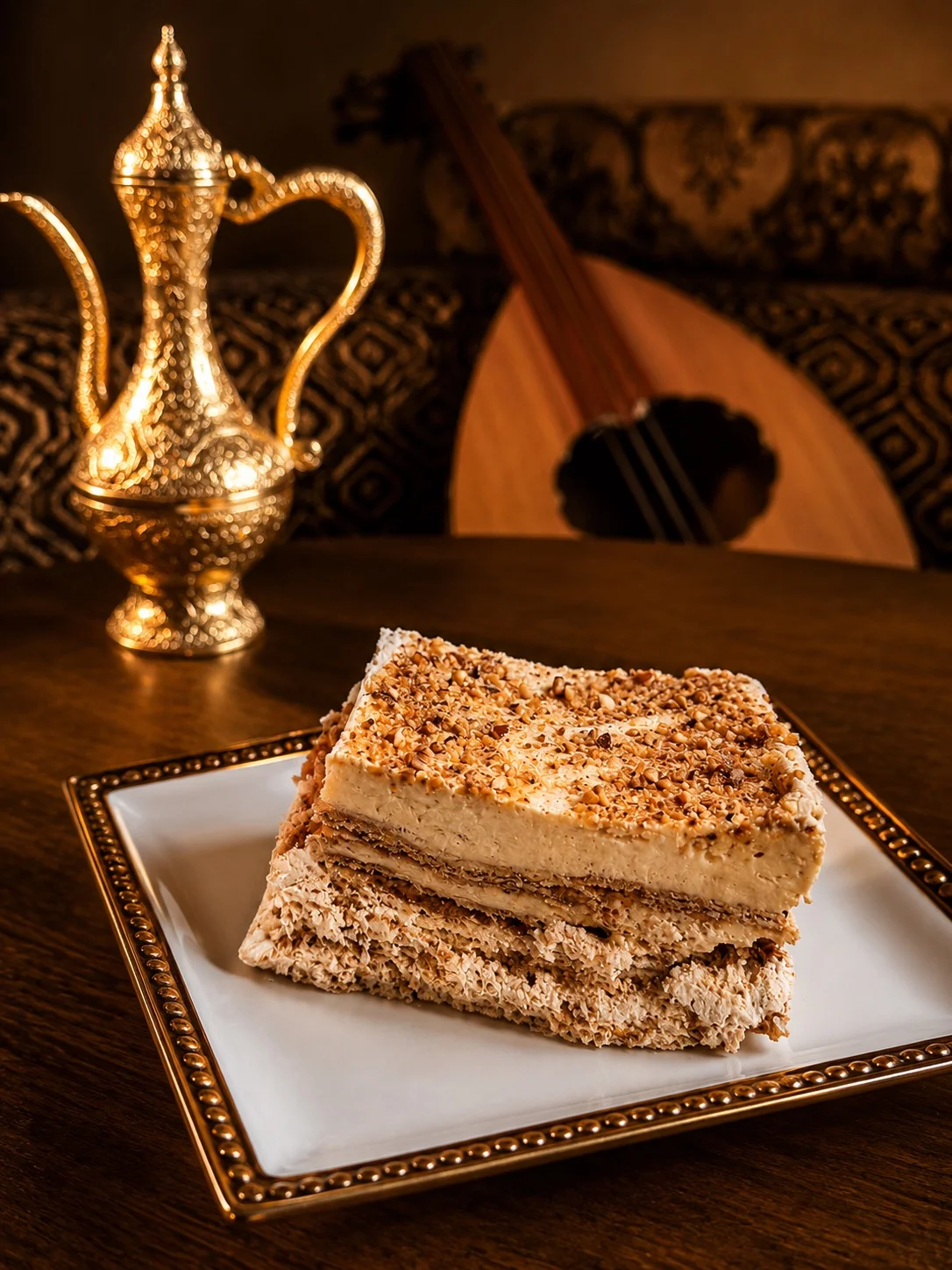 A slice of layered dessert with nuts, a golden coffee pot, and an oud in the background.