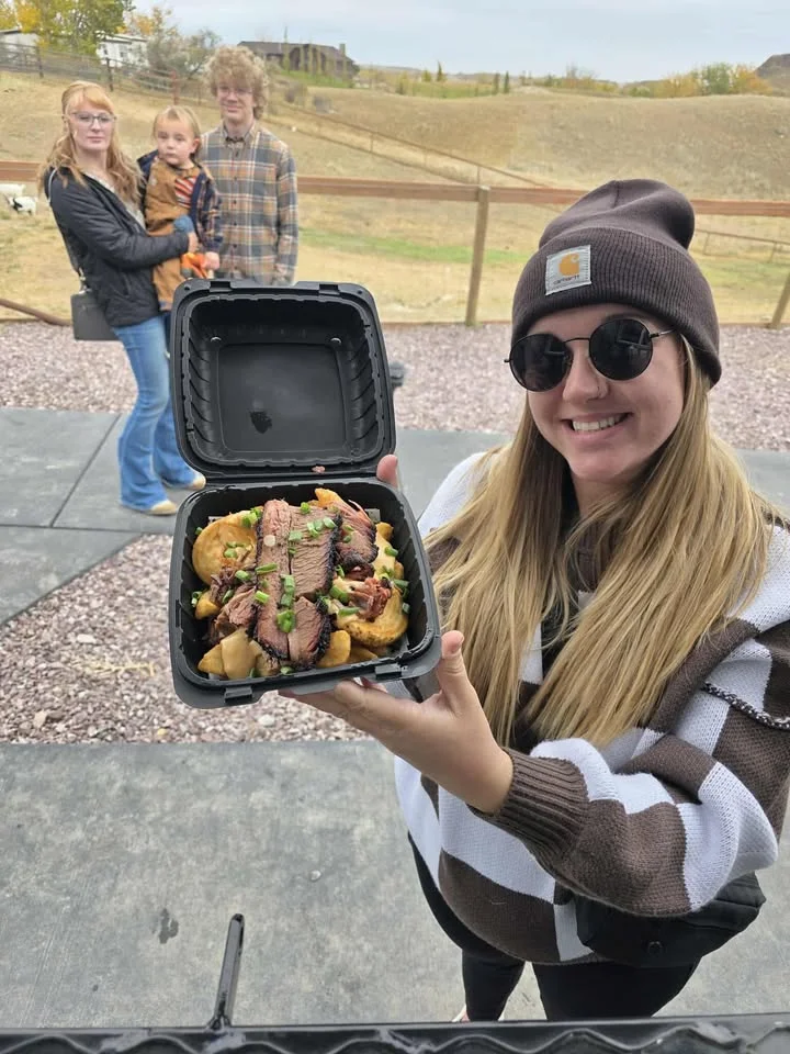 A person smiling and holding a takeout container filled with sliced brisket over a bed of food, with people in the background.