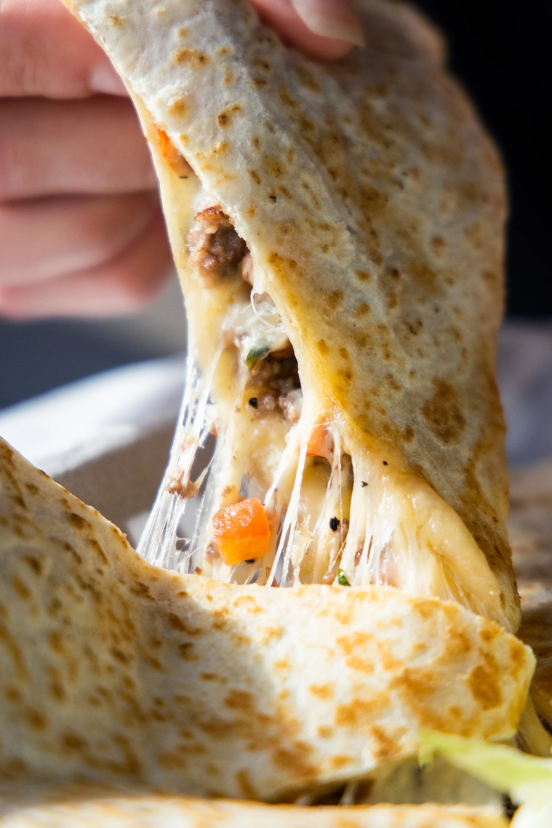 A close-up of a hand pulling apart a cheesy tortilla wrap filled with meat and vegetables.