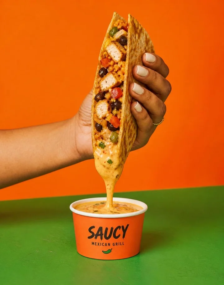 A hand holds a taco filled with ingredients, with cheese sauce dripping into a cup below. The vibrant background adds a lively touch to the scene.