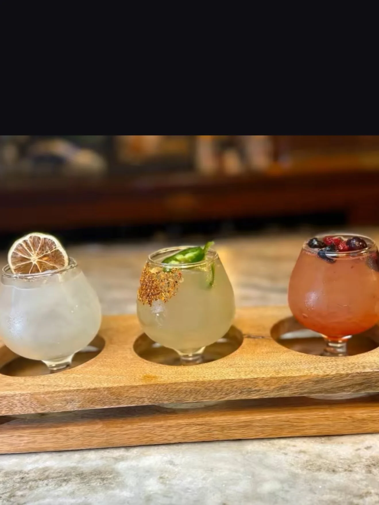 Three colorful cocktails served on a wooden tray, featuring garnishes like lime and a spicy rim.