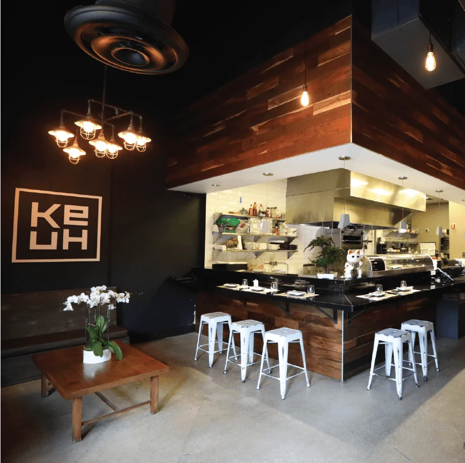 Ke-uH restaurant interior featuring a central wooden sushi bar with white industrial stools, black tables along a brick wall, and warm Edison bulb lighting.