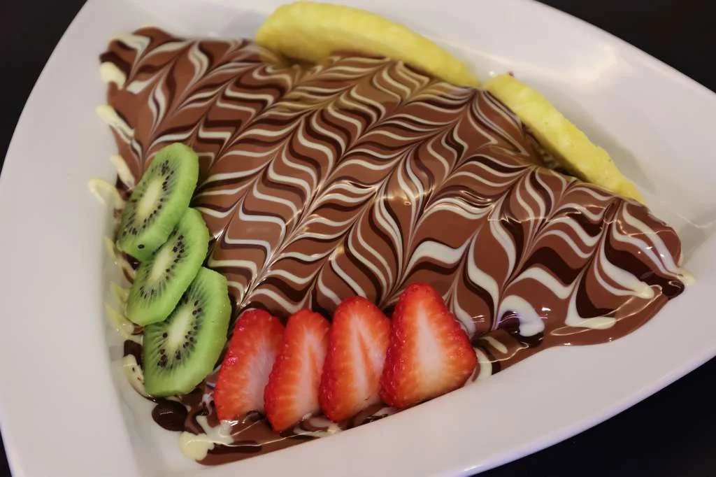 A triangular chocolate dessert garnished with fresh fruits including strawberries and kiwi, and drizzled with chocolate sauce.