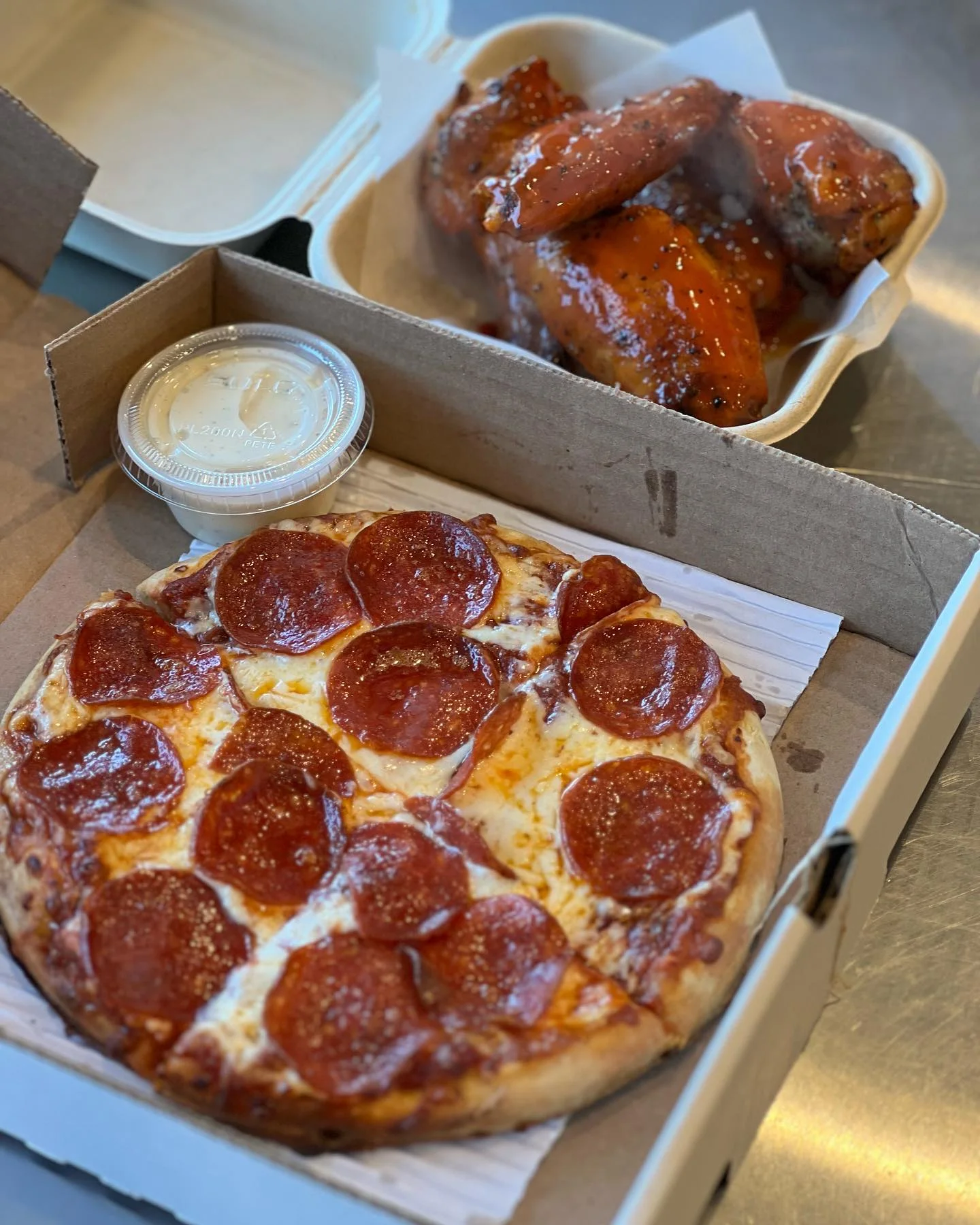 A box containing a pepperoni pizza with a side of chicken wings and a dipping sauce. The pizza features melted cheese and pepperoni slices, while the wings are glazed with a shiny sauce.