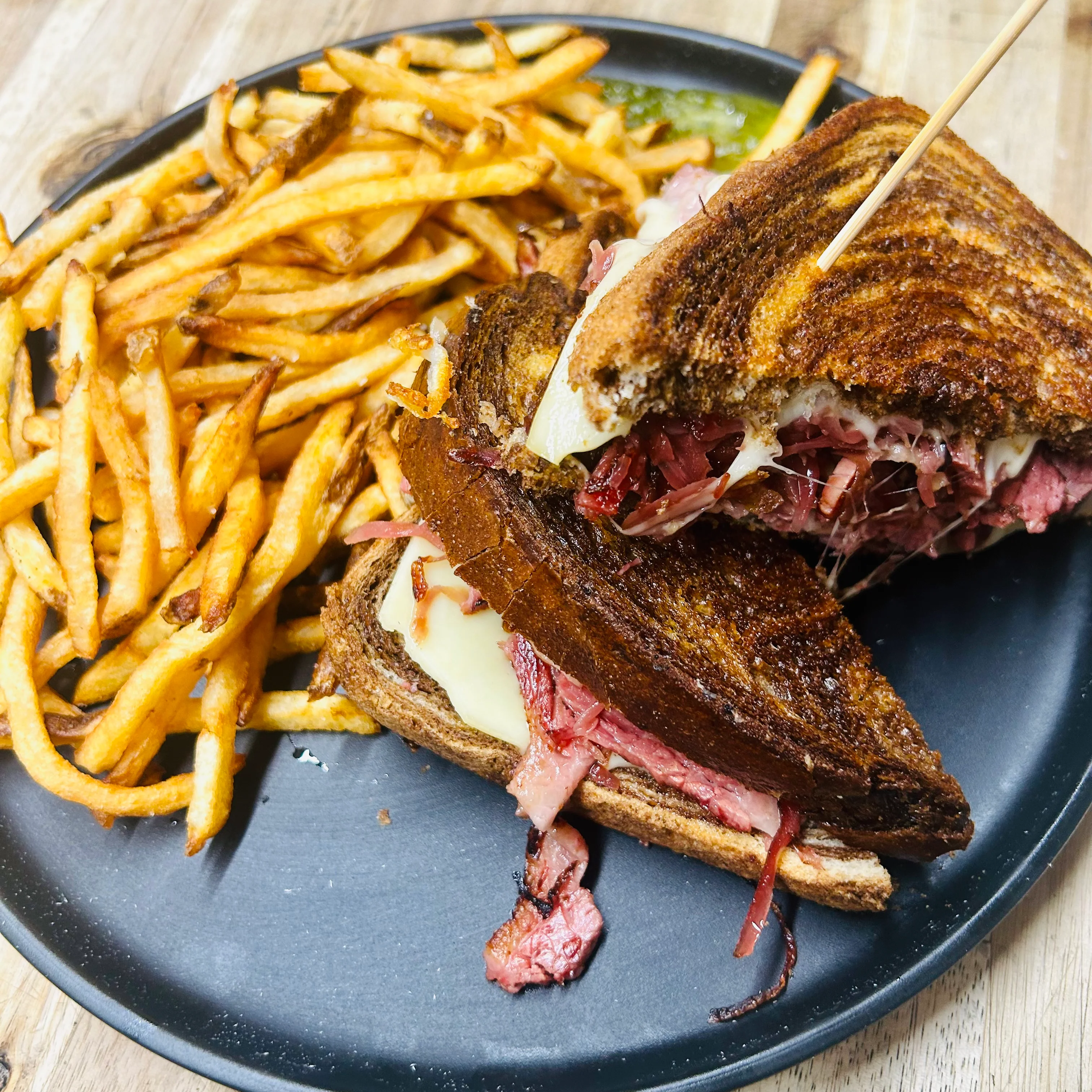 A toasted sandwich filled with pastrami and cheese, served alongside a portion of golden fries on a black plate.
