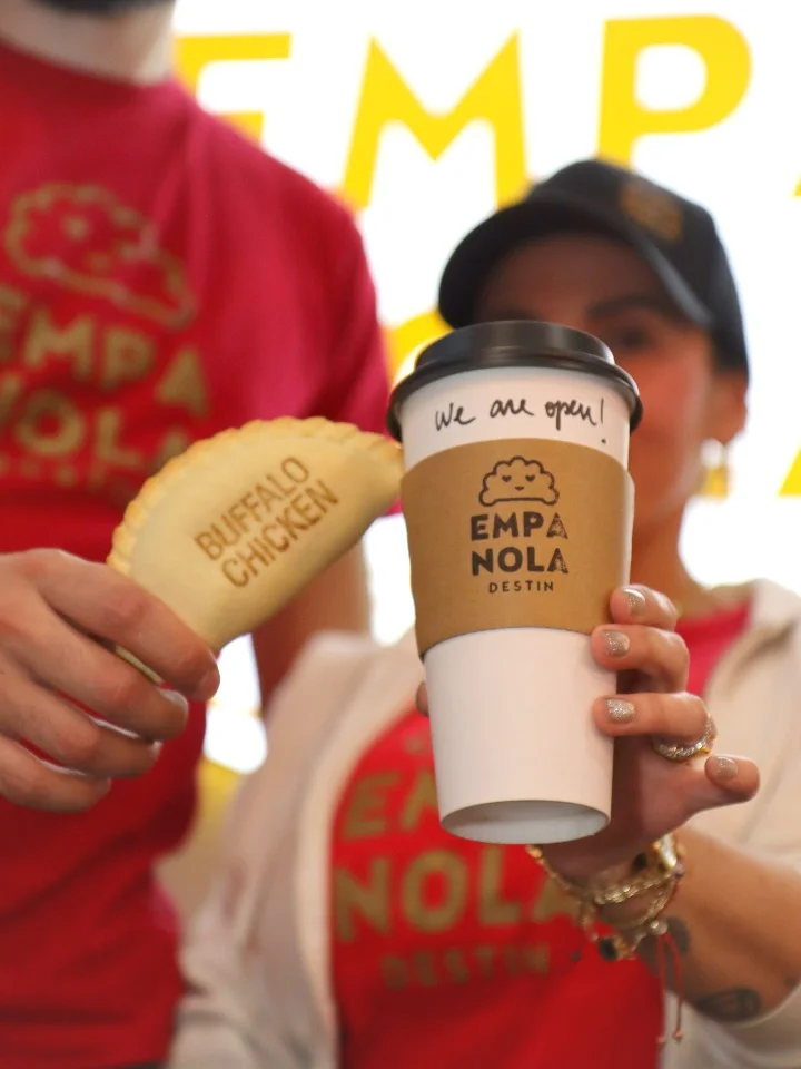 A person holding an empanada with 'Buffalo Chicken' engraved on it, next to a coffee cup with a note saying 'We are open'.