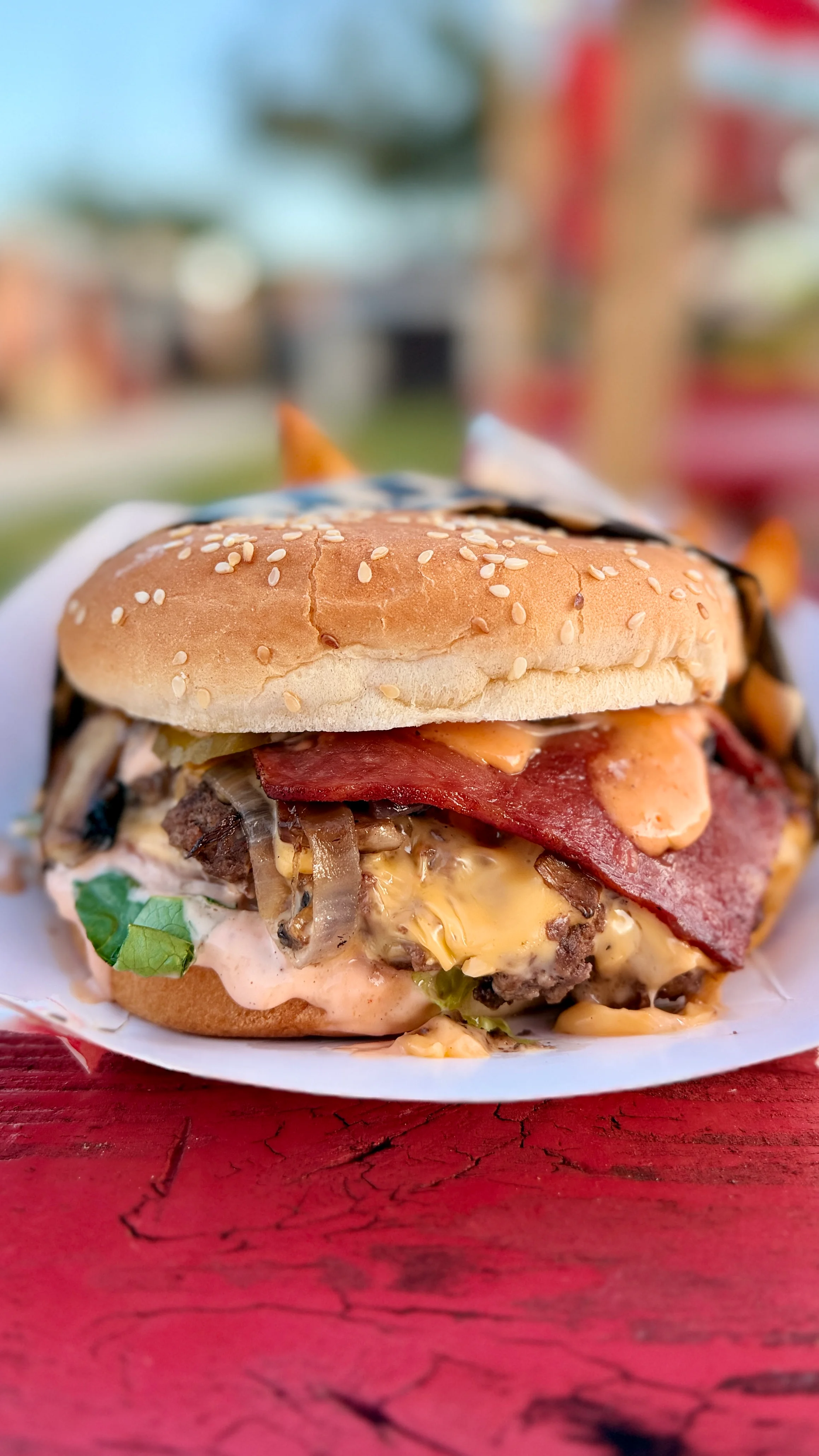 A close-up of a gourmet burger featuring a sesame seed bun, lettuce, cheese, grilled onions, and slices of bacon topped with a special sauce. It is served on a white plate against a rustic red surface.