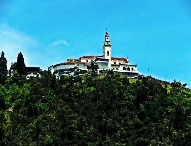 A historic building set atop a hill surrounded by greenery under a clear blue sky, featuring a tall spire and traditional architecture.