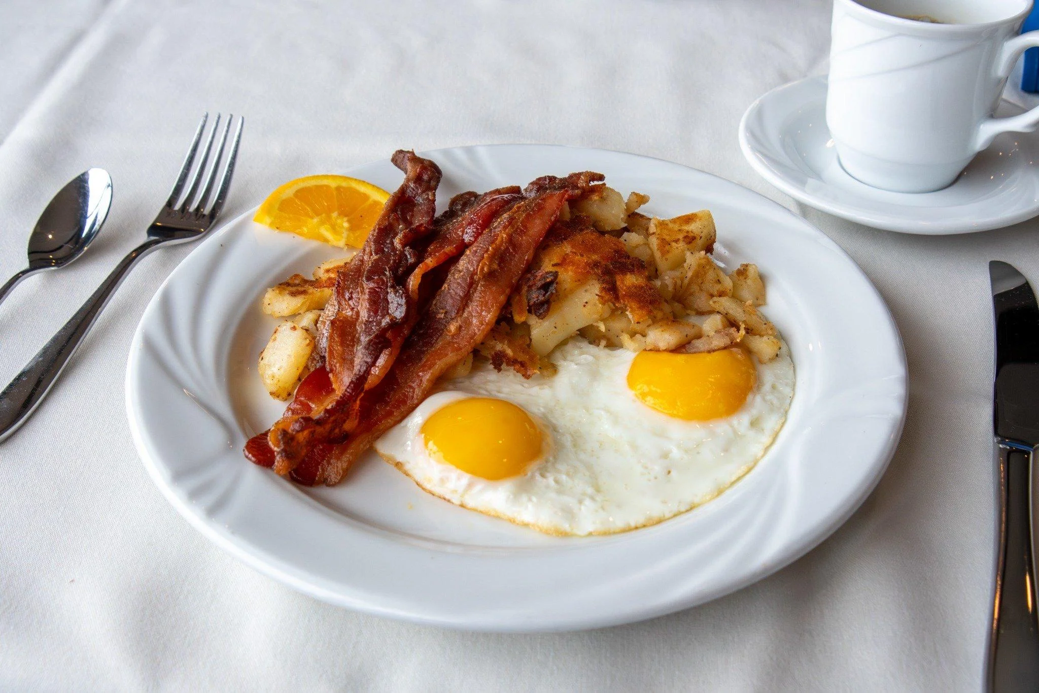 A plate featuring two sunny-side-up eggs, crispy bacon, and golden-brown hash browns, accompanied by an orange slice.